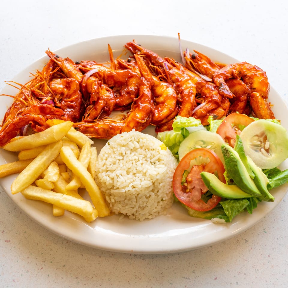 Plate of saucy grilled shrimp served with rice, French fries, and a fresh lettuce, tomato, cucumber, and avocado salad.