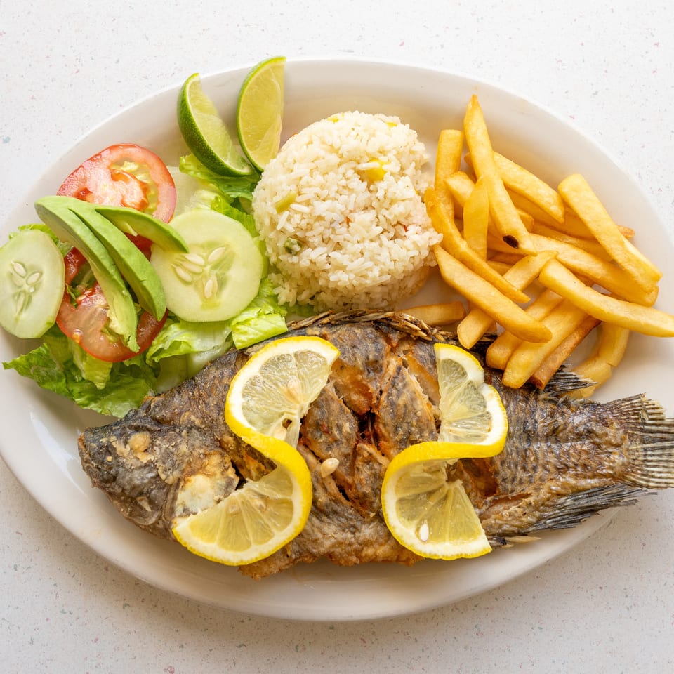 Whole fried fish with lemon slices, rice, French fries, avocado, cucumber, tomato, lettuce, and lime wedges on a platter.