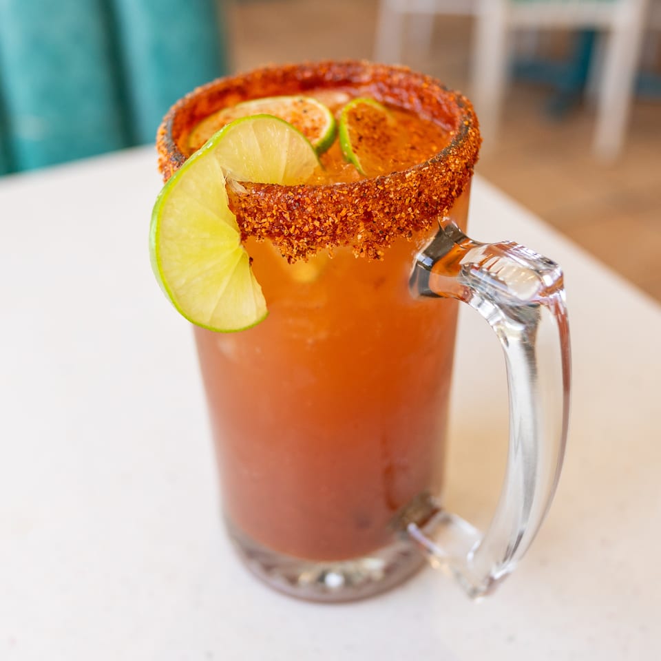 Orange cocktail in a glass mug with a chili-seasoned rim and lime slices, resting on a white table.