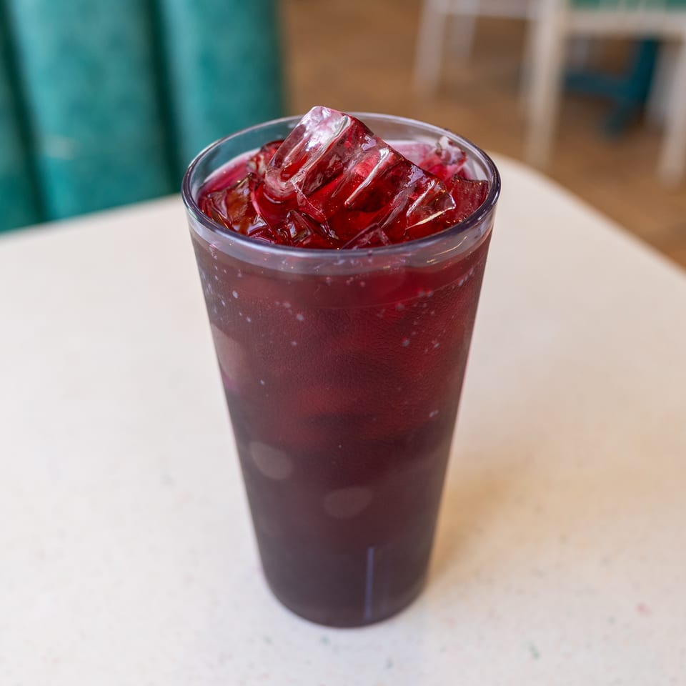 A clear glass holds a dark red iced beverage on a light restaurant table, with teal seating and chairs softly blurred behind it.