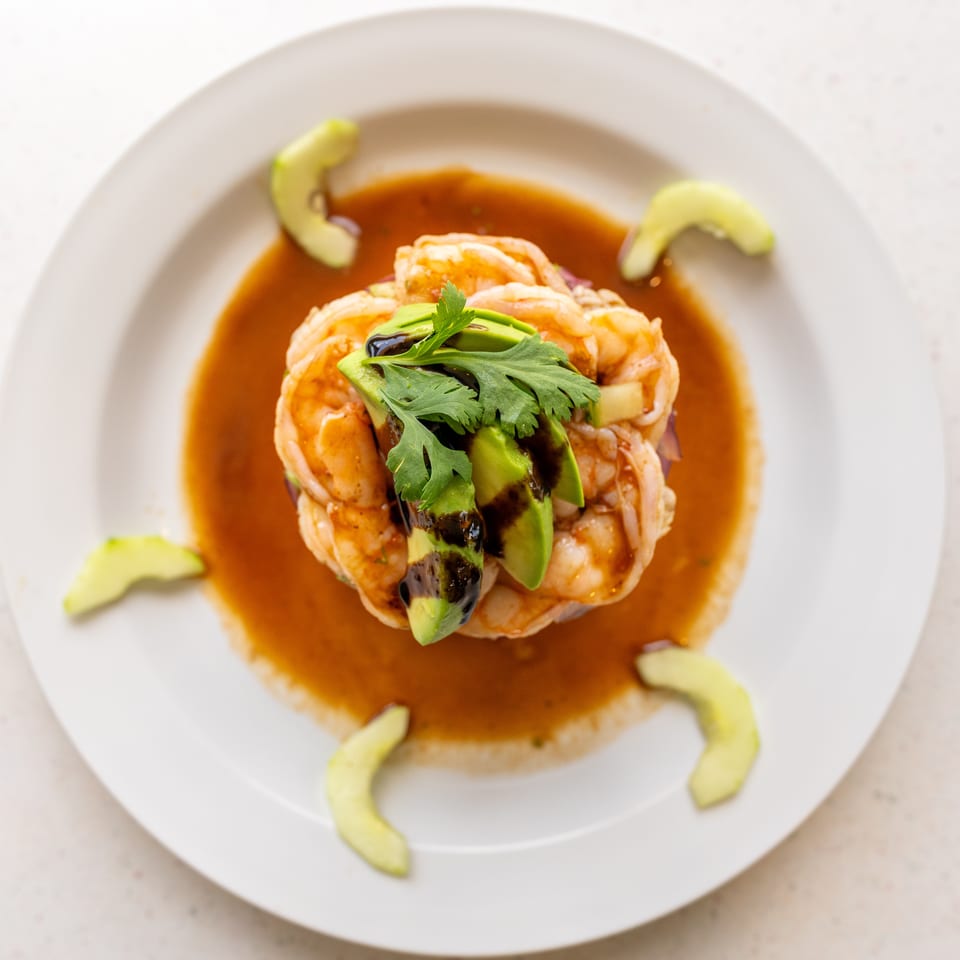 Shrimp arranged around avocado slices and cilantro, served in a rich brown sauce on a white plate.