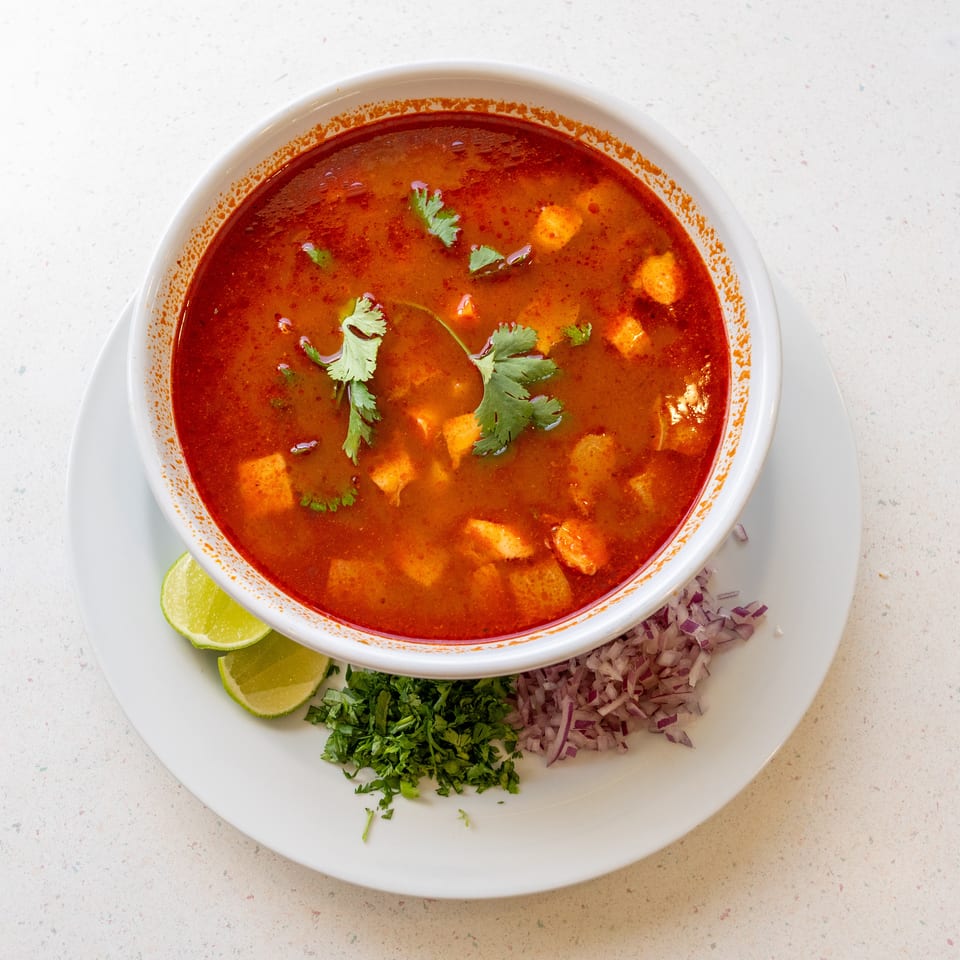 Bowl of red spicy soup garnished with cilantro, served with lime wedges, chopped onion, and herbs.