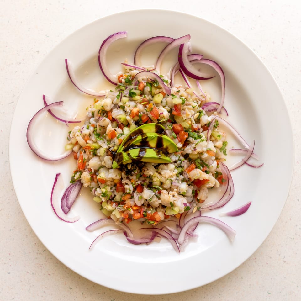 Colorful seafood ceviche topped with avocado and balsamic glaze, garnished with red onion, on a white plate.